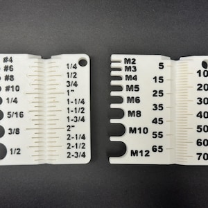 May include: Two white plastic measurement tools with black markings. One tool has fractional inch measurements and drill bit sizes. The other has metric measurements and millimeter increments. Both have a hole for hanging.