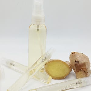 May include: A clear glass spray bottle and several small glass vials filled with a light yellow liquid. Slices of fresh ginger root are also present. The items are arranged on a white surface.