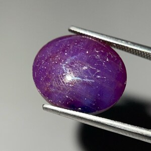 Könnte beinhalten: Ein polierter, oval geformter, violetter Edelstein, der von einer Pinzette gehalten wird. Der Edelstein hat einen sternartigen Effekt mit weißen Linien auf seiner Oberfläche. Der Edelstein ist tiefviolett mit blauen Farbtönen und vor neutralem Hintergrund.