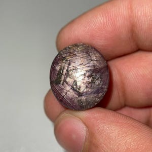 Puede incluir: Una gema redonda y pulida, de un tono púrpura intenso, entrecruzada por finas líneas claras. La piedra tiene una textura ligeramente rugosa y parece estar sujeta en una mano. La gema es un corte cabujón.