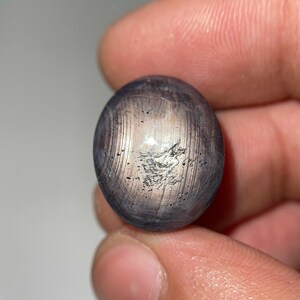 May include: A polished, oval-shaped gemstone with a mix of gray and silver tones. The stone has a reflective surface with visible linear patterns and a subtle, light-colored center. The gemstone is held in a hand, showcasing its natural beauty and unique texture.
