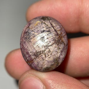 May include: A polished, oval-shaped gemstone with a mix of purple, brown, and white hues. The stone has a network of fine lines across its surface, creating a unique pattern. The gemstone is held between fingers.