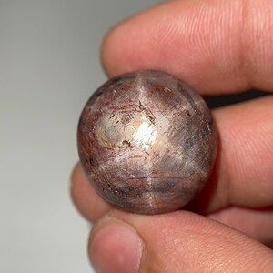 Puede incluir: Una gema esférica pulida con un patrón en forma de estrella. La piedra tiene una mezcla de tonos marrón rojizo y plateado, con líneas y texturas visibles en su superficie. La gema se sostiene en una mano.