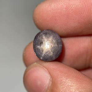 Puede incluir: Una gema de zafiro estrella redonda y púrpura oscuro. La piedra tiene un patrón de estrella de seis rayos en el centro, con una superficie pulida y reflectante. La gema se sostiene entre los dedos.