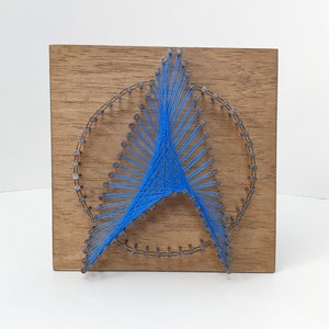 May include: A blue string art piece in the shape of a peace sign on a wooden background.