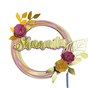 Könnte beinhalten: Ein rosa-goldener Tortendekor mit dem Namen "Shamantha" in goldenen Buchstaben. Der Dekor hat einen Kreis mit rosa-goldenen Streifen, umgeben von rosa und gelben Papierblumen und goldenen Blättern.