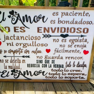 May include: Wooden framed sign with the words "El Amor" in a decorative font. The sign features a white background with black and brown text, including hearts and an arrow design. The text is in Spanish.