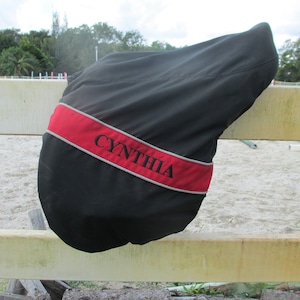 May include: Black saddle cover with a red and grey trim. The cover has the name "CYNTHIA" embroidered on it.