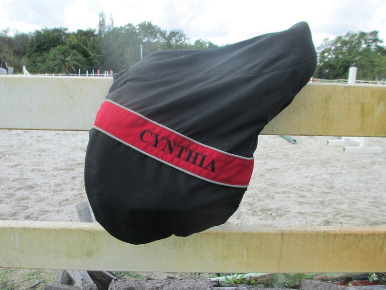 May include: Black saddle cover with a red and grey trim. The cover has the name "CYNTHIA" embroidered on it.