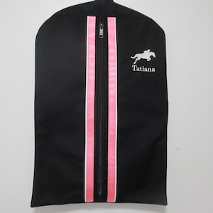 May include: Black garment bag with pink and white stripes down the center. The bag has a zipper closure and the name "Tatiana" embroidered on the front.
