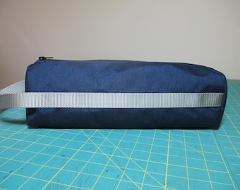 May include: A dark blue zippered pouch with a grey strap. The pouch is lying on a green cutting mat.