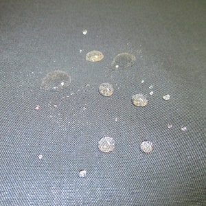 May include: Close-up of a dark grey fabric surface with multiple small water droplets scattered across it.