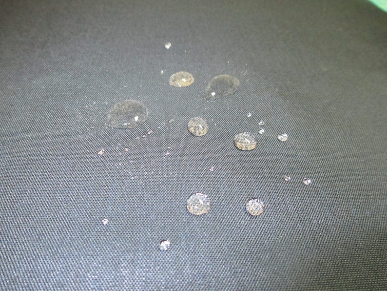 May include: Close-up of a dark grey fabric surface with multiple small water droplets scattered across it.