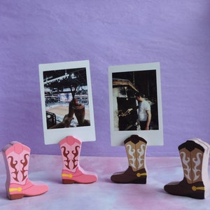 May include: Four pink and brown cowboy boot photo holders with pictures of a baseball game, a person grilling, and a person holding a drink.