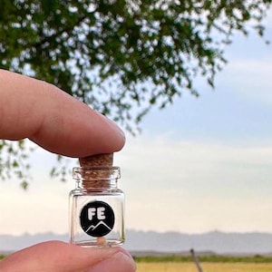 May include: A small glass bottle with a cork stopper. The bottle has a black and white label with the letters "FE" and a mountain range graphic. The bottle is being held by a finger.