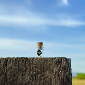 May include: A small glass bottle with a cork top and a black label that says "FAITH". The bottle is sitting on a weathered wooden surface against a blue sky.