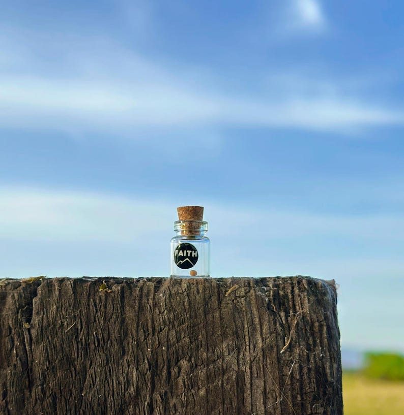 May include: A small glass bottle with a cork top and a black label that says "FAITH". The bottle is sitting on a weathered wooden surface against a blue sky.