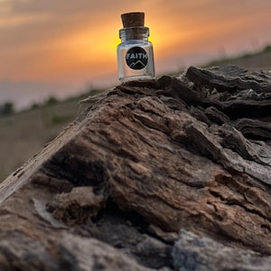 May include: A small glass bottle with a cork stopper and a black label that says "FAITH" on it. The bottle is sitting on a piece of weathered wood with a sunset in the background.