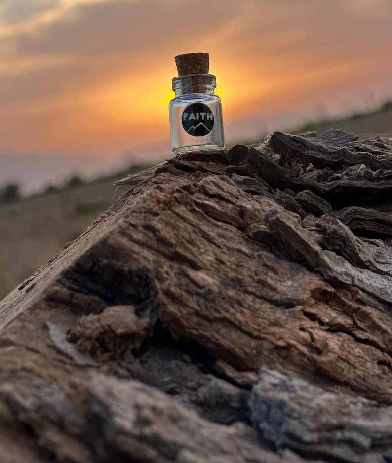 May include: A small glass bottle with a cork stopper and a black label that says "FAITH" on it. The bottle is sitting on a piece of weathered wood with a sunset in the background.