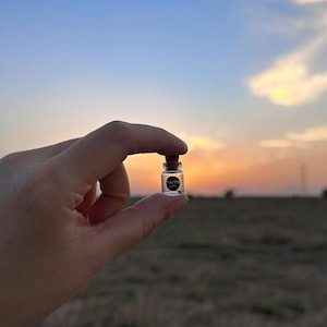 May include: A small glass bottle with a cork stopper, held in a hand against a sunset sky. The bottle has a black label with the word "FAITH" printed on it.