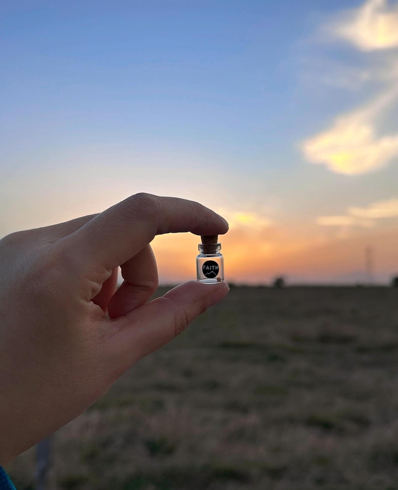 May include: A small glass bottle with a cork stopper, held in a hand against a sunset sky. The bottle has a black label with the word "FAITH" printed on it.