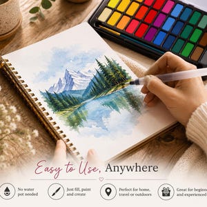 Op de afbeelding: Een spiraalgebonden schetsboek met een aquarel landschapsschildering van besneeuwde bergen, dennenbomen en een meer. Een waterpenseel wordt gebruikt om te schilderen. Bevat een meerkleurige aquarelverfset en de tekst: Easy to Use, Anywhere.