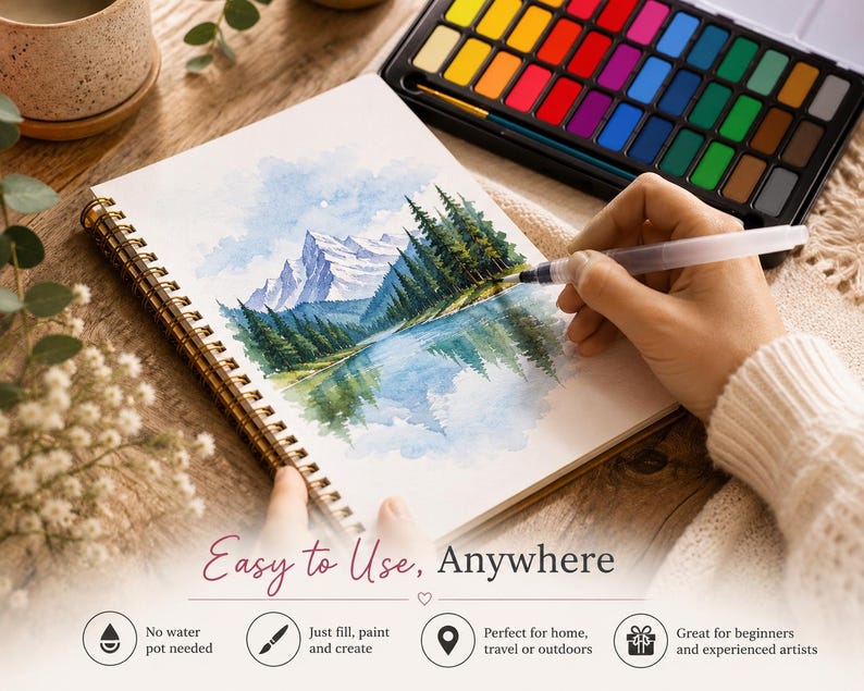 Op de afbeelding: Een spiraalgebonden schetsboek met een aquarel landschapsschildering van besneeuwde bergen, dennenbomen en een meer. Een waterpenseel wordt gebruikt om te schilderen. Bevat een meerkleurige aquarelverfset en de tekst: Easy to Use, Anywhere.