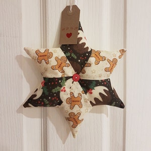 May include: A brown and white star-shaped Christmas decoration with a gingerbread man pattern and a red button in the centre. The decoration is hanging from a white string with a brown tag that says "Spook" with a red heart.