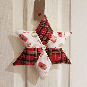 May include: A red and black tartan star-shaped ornament with a white background featuring a coffee cup, candy canes, and other festive designs. The ornament has a button in the centre and a brown tag with the word "Apooh" and a red heart.