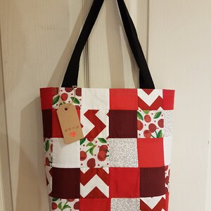 May include: A red and white patchwork tote bag with a cherry print. The bag has black straps and a brown tag that says "MADE WITH LOVE".