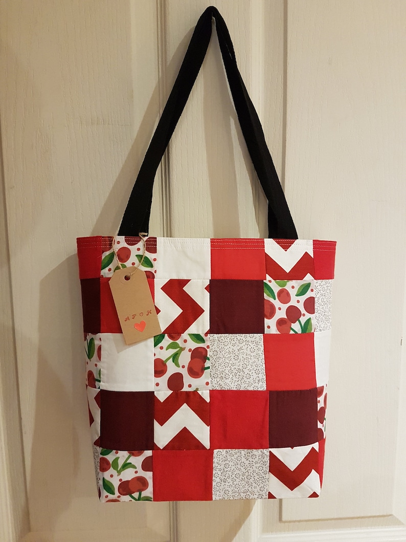 May include: A red and white patchwork tote bag with a cherry print. The bag has black straps and a brown tag that says "MADE WITH LOVE".