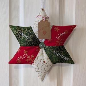 May include: A star-shaped Christmas decoration made of fabric, featuring green, red, and white patterned segments. The ornament has embroidered names and snowflake designs. A small tag with a heart hangs from the top.