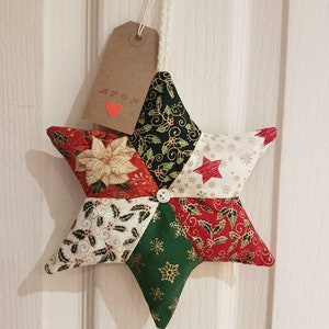 May include: A handmade Christmas star ornament made of patchwork fabric. The star is red, green, white, and gold with a variety of patterns. It has a brown tag with the text "A.P.O.K." and a red heart.