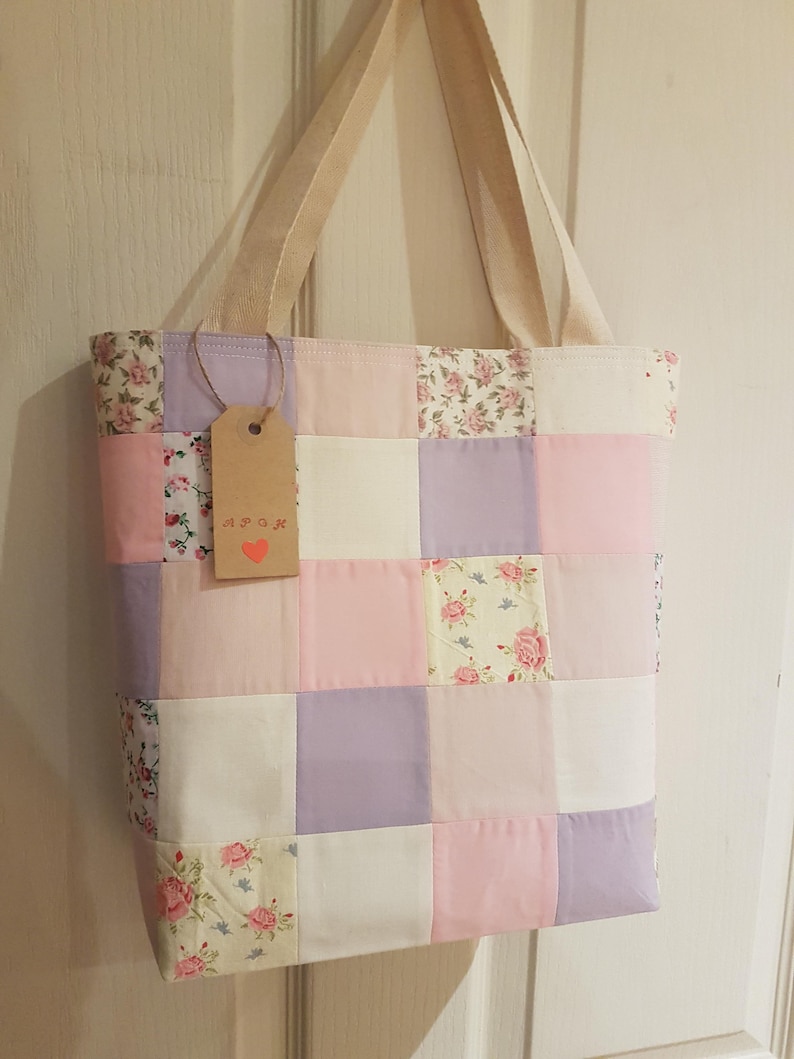 May include: A patchwork tote bag with a cream canvas base and squares of pink, lavender, and floral fabric. The bag has two cream canvas straps and a small brown tag with the word "Spook" written on it.