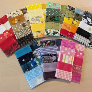 May include: A selection of patchwork fabric wallets in various colours and patterns. Each wallet is made from different fabrics, including floral, polka dot, and geometric designs. They are secured with a button fastening. The wallets are arranged in a fan shape.