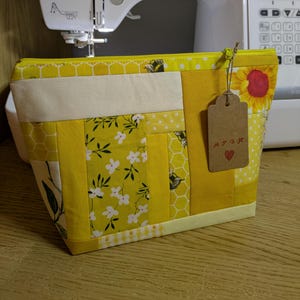 May include: A handmade yellow and white patchwork zippered pouch. The pouch features various fabric patterns, including floral, honeycomb, and bee designs. A brown tag with the letters "APGK" and a heart is attached to the zipper.