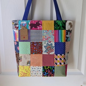 May include: A patchwork tote bag with a variety of colourful fabric squares. The bag has blue handles and a small tag with text and a heart. The bag is hanging on a white door.