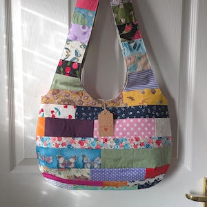 May include: A patchwork shoulder bag with a variety of fabric patterns and colors, including floral, polka dots, and stripes. The bag has a wide strap and a small tag with text and a heart. The bag is hanging on a white door.