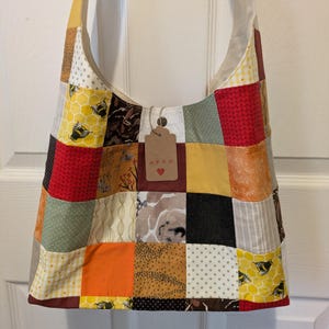 May include: A patchwork tote bag featuring a mix of fabrics in various patterns and colours, such as yellow honeycomb, red knit, and floral designs. The bag has a cream lining and a tag with the text "ASON" and a heart symbol.
