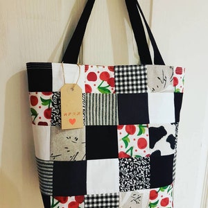 May include: A black and white patchwork tote bag with a cherry print. The bag has black straps and a brown tag with the word "APOK" written on it.