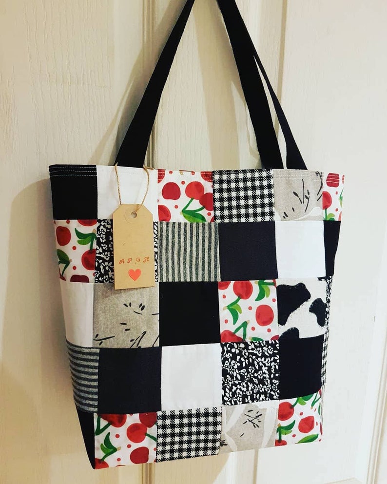 May include: A black and white patchwork tote bag with a cherry print. The bag has black straps and a brown tag with the word "APOK" written on it.