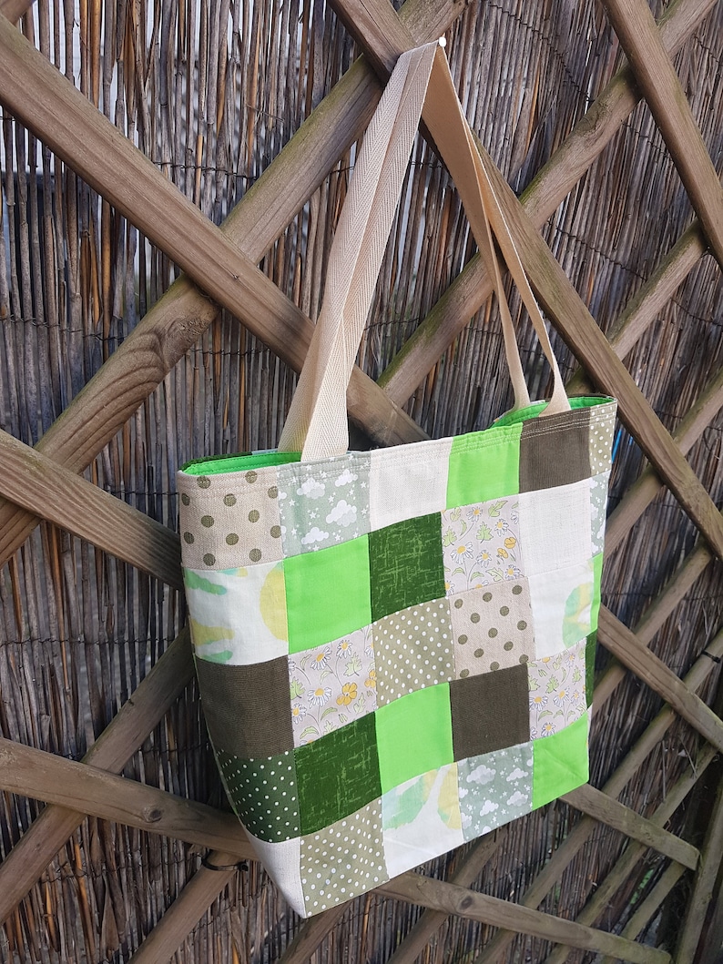 May include: A patchwork tote bag with a green, brown, and beige color scheme. The bag has a long, beige strap and is made of different fabrics with various patterns.