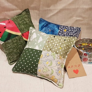 May include: Four patchwork fabric pincushions, one with a watermelon print, one with a green and white polka dot print, one with a floral print, and one with a blue and white print. A tin filled with sewing pins is in the background.