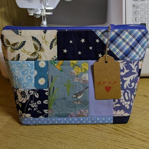 May include: A patchwork make-up bag with a blue zip and a brown tag. The bag features various fabric patterns, including butterflies, stars, and floral designs. The tag has the letters "APGK" and a red heart.