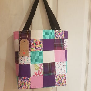 May include: A patchwork tote bag with a variety of colorful fabrics, including purple, pink, white, and green. The bag has black straps and a brown tag with the word "Arox" written on it.