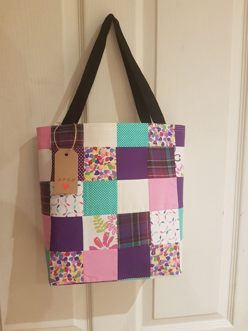 May include: A patchwork tote bag with a variety of colorful fabrics, including purple, pink, white, and green. The bag has black straps and a brown tag with the word "Arox" written on it.