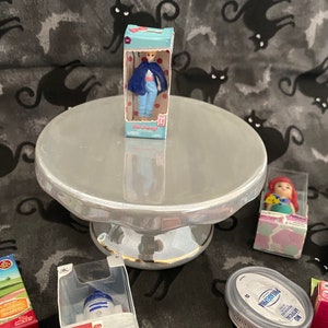 May include: A collection of miniature toys and figurines, including a doll in a blue dress, a robot, a princess, and a mermaid. The toys are displayed on a white cake stand and a black and white patterned surface.