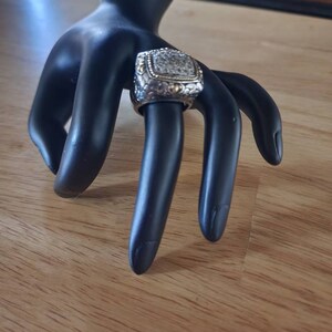 May include: A silver and gold ring with a square-shaped stone set in the center. The stone is surrounded by a decorative border. The ring is on a black hand model.