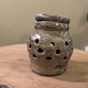 May include: A handcrafted ceramic jar with a lid, featuring a brown and tan mottled glaze. The jar has numerous circular openings around its body. The lid is a darker brown with a textured surface. The jar sits on a wooden surface.