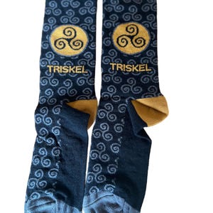 Op de afbeelding: Een paar zwarte sokken met een gouden triskelion-symbool en het woord "TRISKEL" gedrukt op elke sok. De sokken hebben een blauw en grijs swirl-patroon.